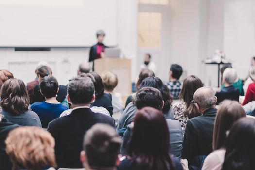 Business and entrepreneurship symposium. Female speaker giving a talk at business meeting. Audience in conference hall. Rear view of unrecognized participant in audience.