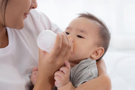 Young asian mother embracing and feeding little baby girl with bottle of milk at home, newborn innocence drinking with mom satisfied, relationship and bonding of mum and child, family concept.