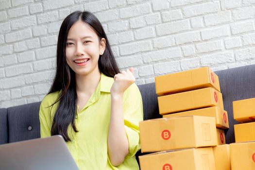 Young asian woman is merchant using laptop computer on sofa checking order of customer with purchase online shopping, business SME or startup, entrepreneur or seller and service delivery product.
