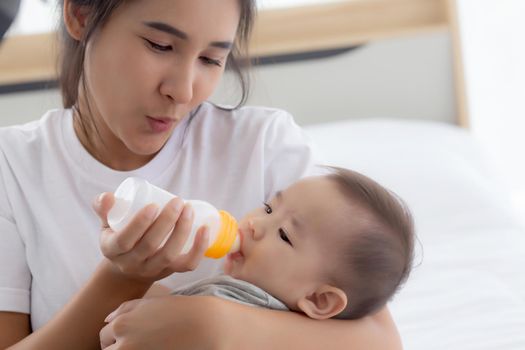 Young asian mother embracing and feeding little baby girl with bottle of milk at home, newborn innocence drinking with mom satisfied, relationship and bonding of mum and child, family concept.