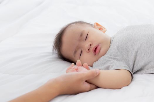 Young asian mother touch hand of little baby girl with tender on bed in the bedroom, mom love newborn and care, woman with expression with child together, parent and daughter, family concept.