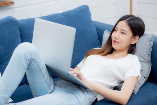 Young asian business woman smile and work from home with laptop computer online to internet on sofa in living room, freelance girl using notebook on couch with comfort, new normal, lifestyle concept.