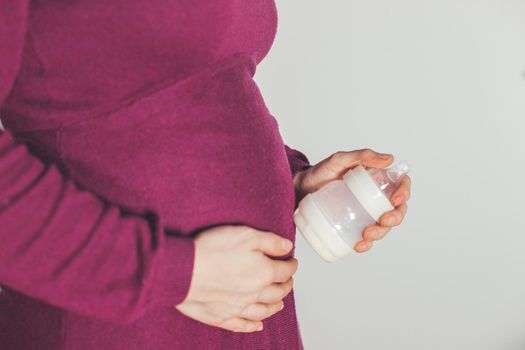Close up of young mother holding a baby bottle on her pregnant tummy, Caucasian, magenta dress