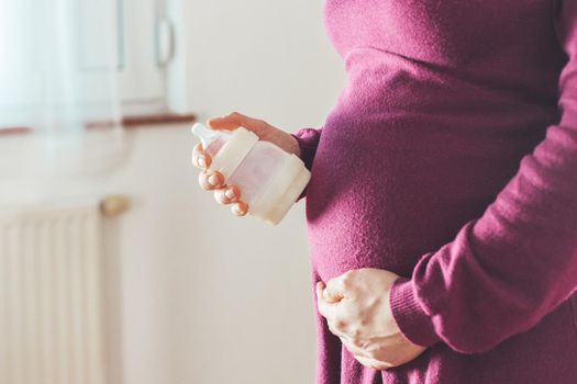 Close up of young mother holding a baby bottle on her pregnant tummy, Caucasian, magenta dress