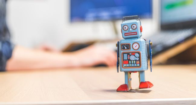 Toy robot on a desktop, man working on the computer in the background. Symbol for artificial intelligence, chatbots or social bots and algorithms