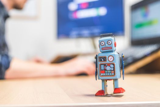 Toy robot on a desktop, man working on the computer in the background. Symbol for artificial intelligence, chatbots or social bots and algorithms