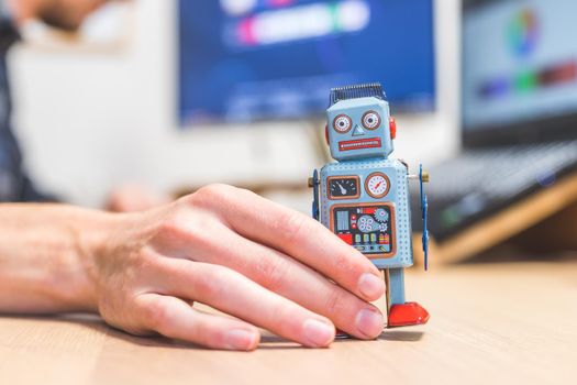 Toy robot on a desktop, man working on the computer in the background. Symbol for artificial intelligence, chatbots or social bots and algorithms