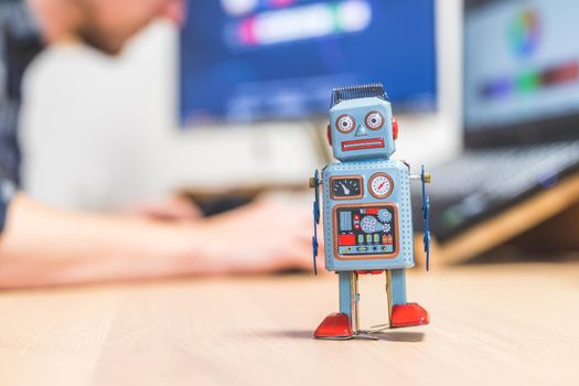 Toy robot on a desktop, man working on the computer in the background. Symbol for artificial intelligence, chatbots or social bots and algorithms