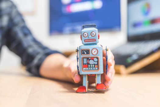 Toy robot on a desktop, man working on the computer in the background. Symbol for artificial intelligence, chatbots or social bots and algorithms