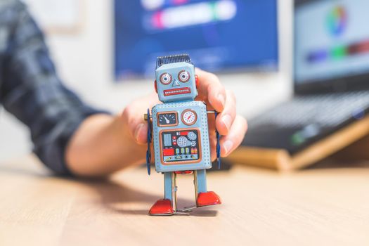 Toy robot on a desktop, man working on the computer in the background. Symbol for artificial intelligence, chatbots or social bots and algorithms