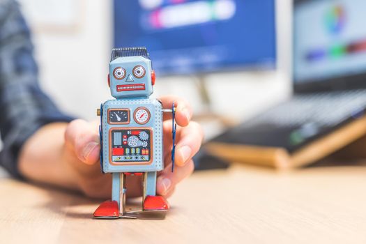 Toy robot on a desktop, man working on the computer in the background. Symbol for artificial intelligence, chatbots or social bots and algorithms