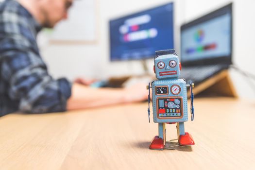 Toy robot on a desktop, man working on the computer in the background. Symbol for artificial intelligence, chatbots or social bots and algorithms