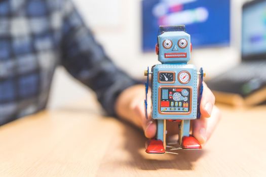 Toy robot on a desktop, man working on the computer in the background. Symbol for artificial intelligence, chatbots or social bots and algorithms
