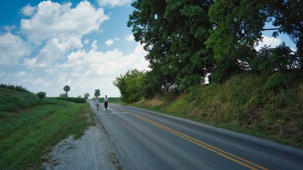 Two Amish Teenagers One Boy and One Girl on Scooters on a Road on a Summer Day