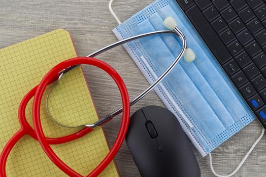 Virus pandemic and medical concept with a colorful red stethoscope coiled over a surgical face mask alongside a notebook on a wooden desk with computer keyboard viewed top down
