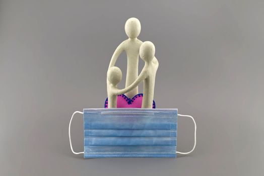 Covid-19 or coronavirus family concept with a small figurine of parents and a child embracing with pink heart and protective surgical face mask in the foreground over a grey background
