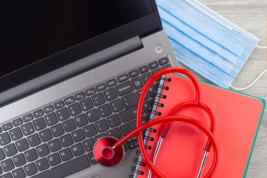 Virus pandemic and medical concept with a colorful red stethoscope coiled over a surgical face mask alongside a notebook on a wooden desk with computer keyboard viewed top down