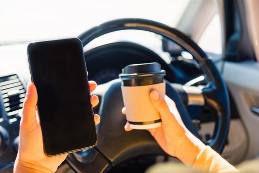 Asian woman drinking hot coffee takeaway cup inside a car and using smartphone blank screen while driving the car in the morning during going to work on highway, Transportation and vehicle concept