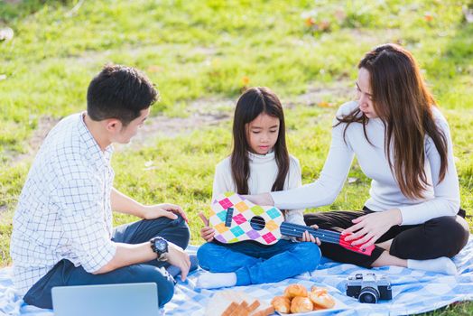 Happy Asian young family father, mother and children having fun and enjoying outdoor together sitting on the grass party with playing Ukulele during a picnic in the garden park on a sunny day
