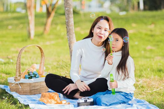 Happy Asian mother and little girl daughter child having fun and enjoying outdoor together sitting on the grass blowing soap bubbles during a picnic in the garden park on a sunny day family time