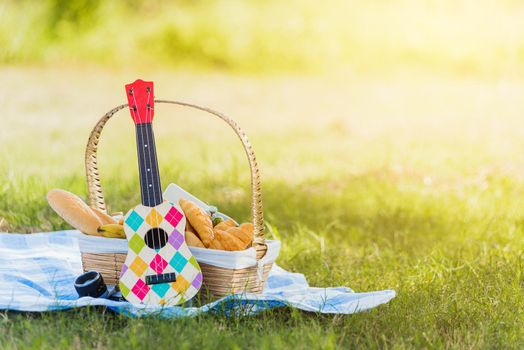 Picnic wattled basket with bread food and fruit, Ukulele, a retro camera on blue cloth in green grass garden with copy space at sunny summertime