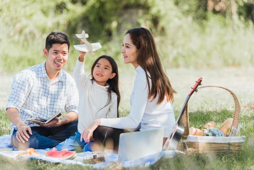 Happy Asian young family father, mother and child little girl having fun and enjoying outdoor sitting on picnic blanket playing toy aircraft at summer garden park