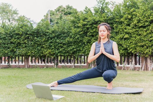 Asian young woman using laptop during online video training practicing yoga outdoors in meditate pose sitting on green grass in nature a field garden park, Stretching health care concept