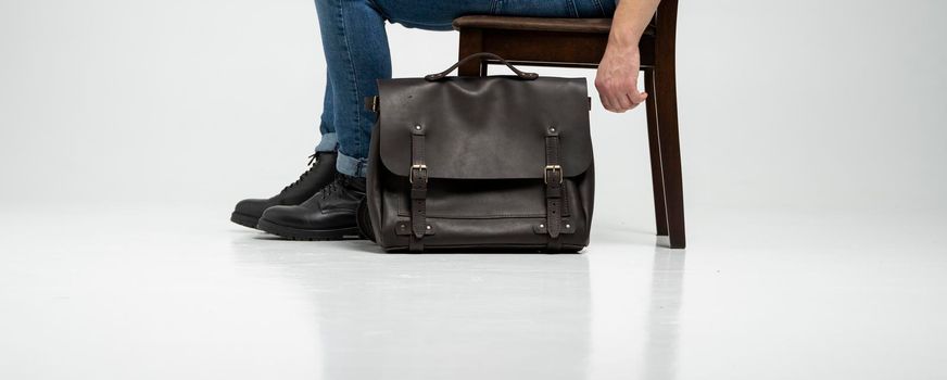 Man in a blue jeans and black boots sits on a chair with a brown men's shoulder leather bag for a documents and laptop on a white floor. Mens leather satchel, messenger bags, handmade briefcase