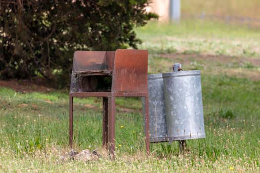 An old rusty BBQ and rubbish bin in a green field with trees and weeds