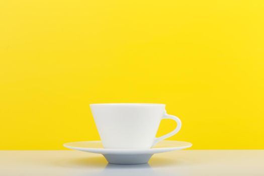 Minimalistic still life with white glossy cup of coffee on white table against yellow background with copy space. High quality photo