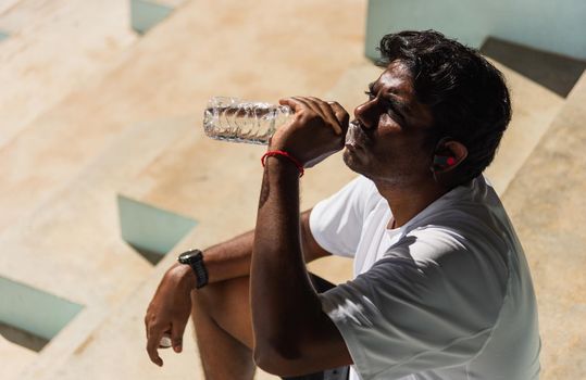 Close up Asian young sport runner black man wear athlete headphones he drinking water from a bottle after running at the outdoor street health park, healthy exercise workout concept