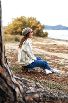 Beautiful Caucasian young woman sitting near the water on a tree trunk in pine forest enjoying the magic winter sea. Happy female person lives the embrace of nature without needing anything else