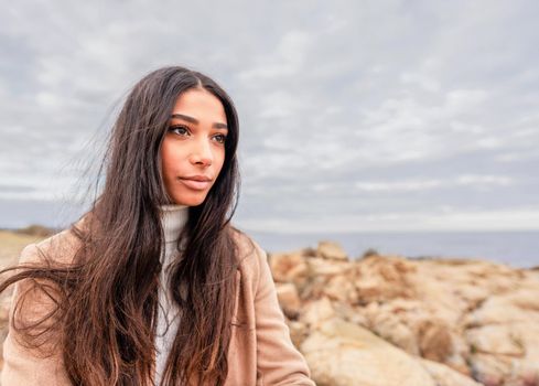 Close up portrait of young beautiful Hispanic woman with long dark hair sitting on sea rocks dreaming looking to the horizon with dramatic grey sky. Fall-winter color mood photography with copy space