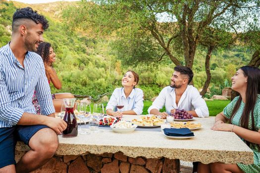 Group of young mixed-race people having fun outdoor in the countryside eating snacks and drinking red wine for an aperitif. Beautiful happy friends bonding smiling each other sitting at a laden table