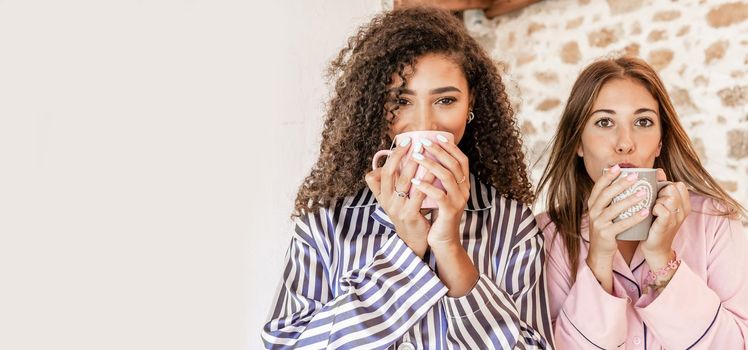 Female beautiful multiracial couple in pajama looking at the camera holding and drinking a cup of tea. Hispanic curly woman and her Caucasian girlfriend posing for glamour photography with a mug