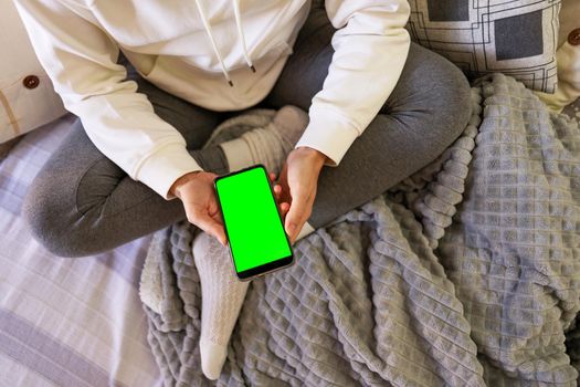 Top view of young woman sitting on the couch wearing comfortable clothes with the smartphone in hand. Female person sitting with crossed legs holds the cellphone with blank green screen for copy space