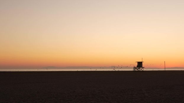 Summertime travel concept. Dark silhouette, iconic retro wooden lifeguard watch tower against sunset orange sky. Contrast watchtower outline, california pacific ocean beach twilight aesthetic, USA.