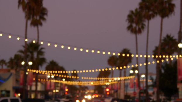 Decorative staring garland lights, palm trees silhouettes, evening sky. Blurred Background. Street decorated with lamps in California. Festive illuminations, beach party, tropical vacations concept