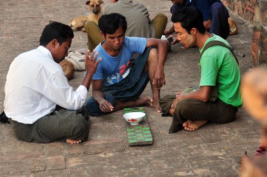 BAGAN, MYANMAR - NOVEMBER 18, 2015: Poor men playing game on street, Side view of group of poor barefoot men sitting on pavement playing oriental game. Portrait Mingalazedi Sulamani Shwezigon Ananda