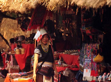 CHIANG MAI, THAILAND - MARCH 1, 2013: Young woman at stall in oriental village wearing traditional gown and standing at stall selling colorful authentic souvenirs. Noth tribes Akha, Lisu, Karen, Lahu