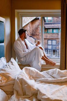 man looking out the window in the morning just wake up with coffee during lockdown corona pandemic covid 19, young men in bathrobe sitting in window daytime