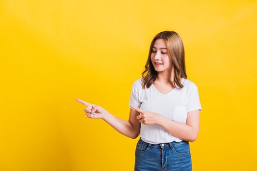 Asian Thai happy portrait beautiful cute young woman standing wear white t-shirt pointing finger away side looking to away side, studio shot isolated on yellow background with copy space