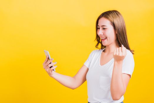 Asian Thai portrait happy beautiful cute young woman smiling stand wear t-shirt lift hand celebrating winning with smart mobile phone looking the phone isolated, on yellow background with copy space