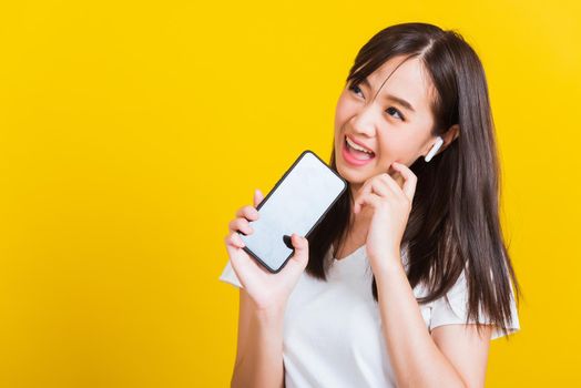 Portrait Asian of a happy beautiful young woman holding a mobile smart phone and wearing wireless headphones listening to music from smartphone studio shot isolated on yellow background