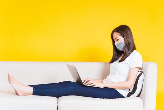 Portrait Asian of beautiful young woman sitting on sofa wearing medical face mask protective she work from home with laptop computer during Coronavirus studio shot isolated on yellow background