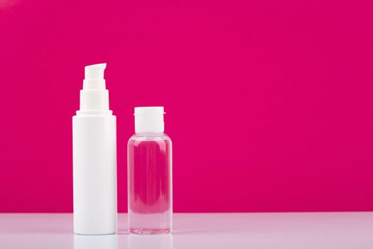 Still life with two cosmetic products, face cream in white tube and skin lotion in transparent bottle on white table against pink background with copy space. High quality photo