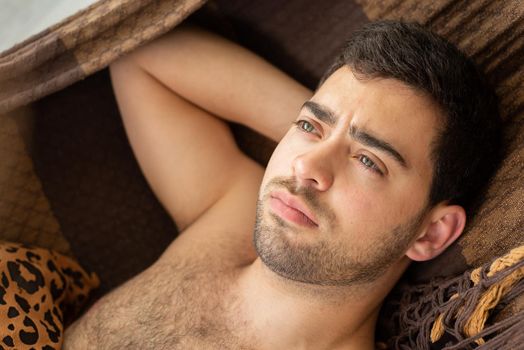 Dark haired bare chested unshaven young man resting in a brown hammock.