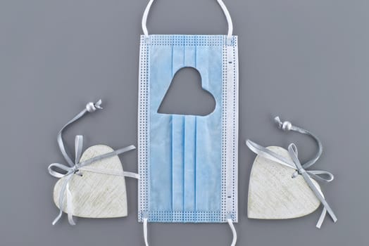 Two hearts between a medical face mask with cut out heart shape on a gray background. Social distancing and contact-less greetings