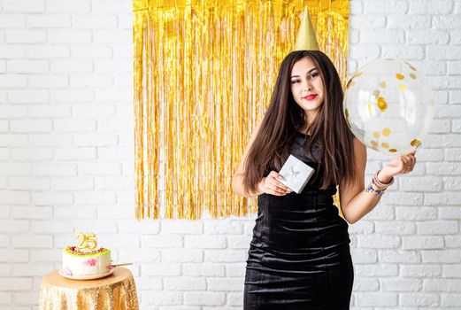 Birthday party. BBeautiful young woman in party dress and birthday hat holding balloon on golden background