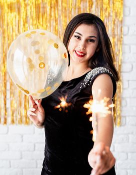 Birthday party. Beautiful young woman holding sparkler and balloon on golden background